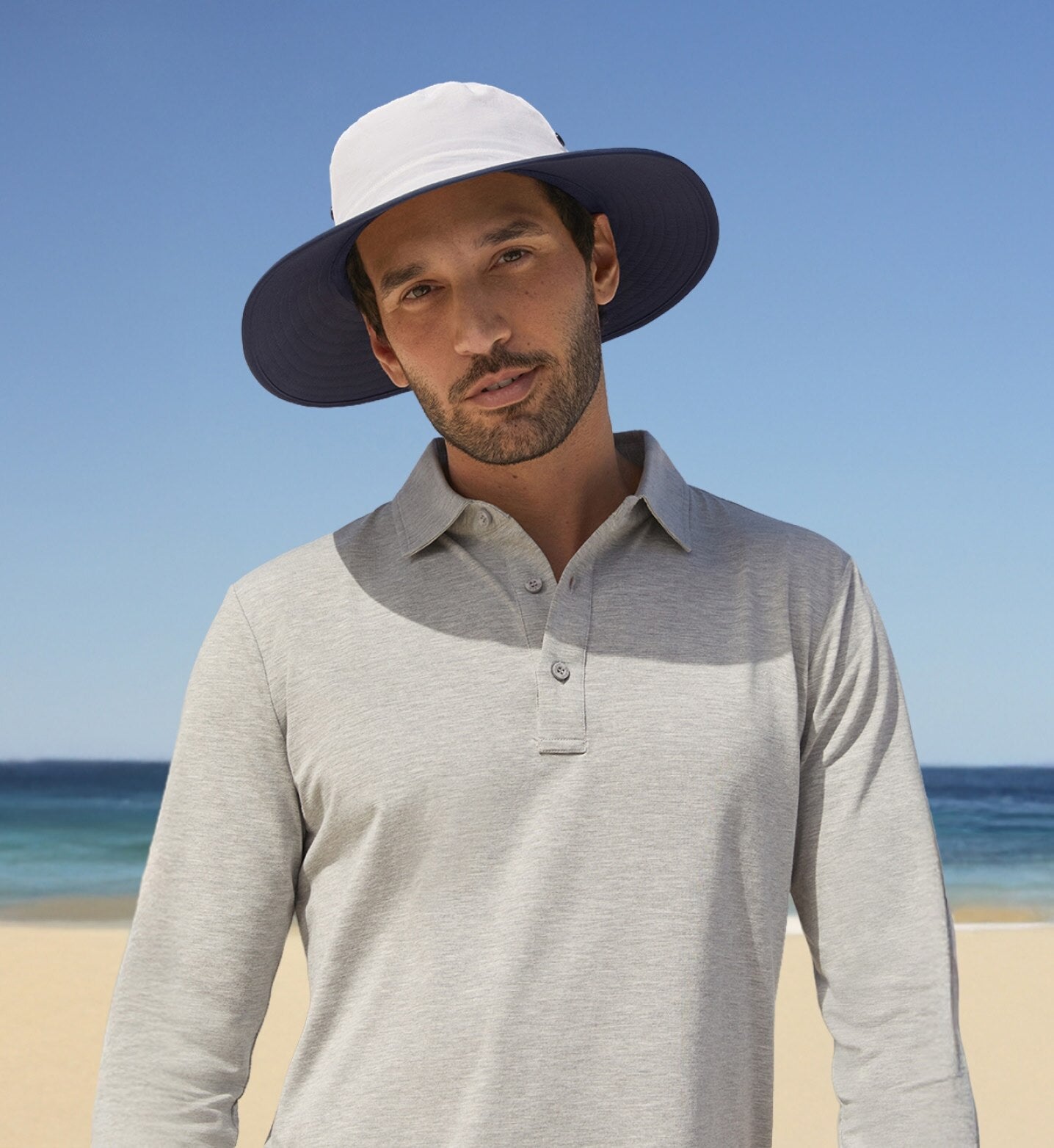 Man wearing a wide-brimmed hat on a beach with clear blue sky and ocean.